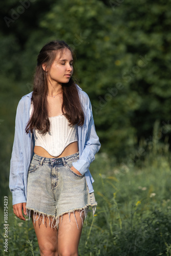 Young woman in casual summer outfit walks along a forest path surrounded by lush greenery and tall trees, sunlight filtering through the leaves