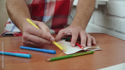 Male hand colors paper craft on table with pencil close up. Decorating and drawing on origami using shading techniques with colored pencils.