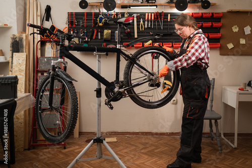Professional female mechanic repairing bicycle in workshop with tools on wall