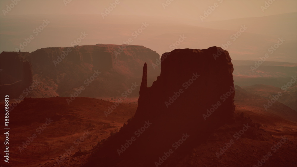 Fototapeta premium The sun sets behind impressive rock formations in a vast desert landscape, creating an enchanting atmosphere with hues of orange and brown. Shadows lengthen as night approaches.