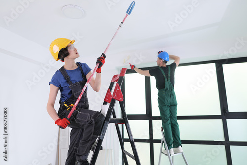 A woman stands on a ladder, expertly applying paint to the ceiling, while a man works on another ladder, adding finishing touches in a contemporary room filled with natural light.