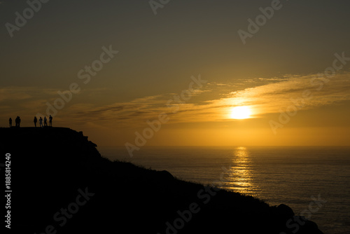 Group of friends silhouetted against a golden sunset sky on a cliff edge in Galicia. Panoramic coastal view
