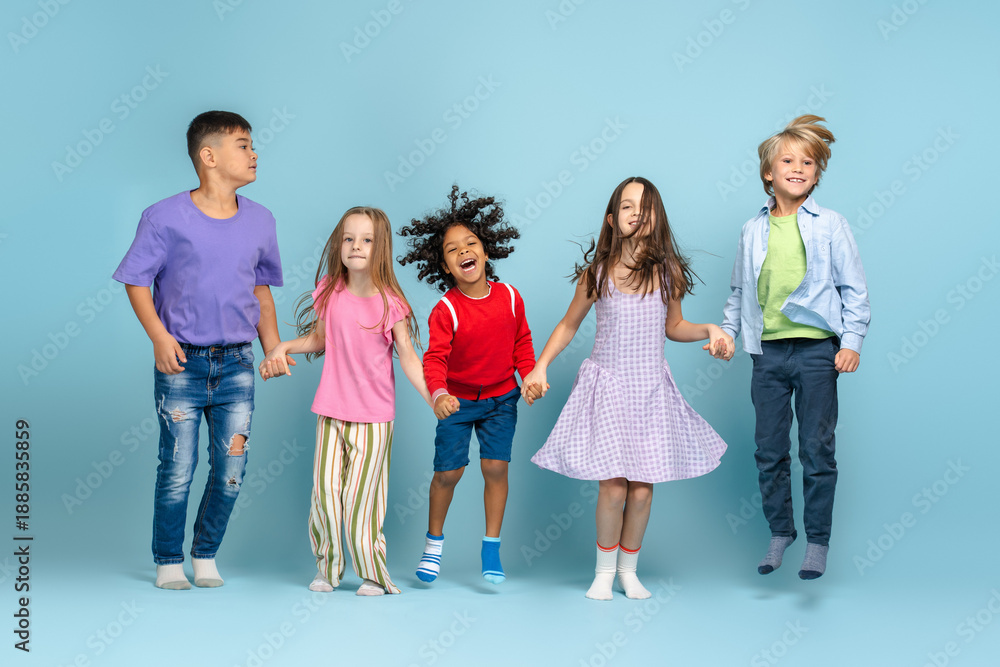 Fototapeta premium Group of positive diverse children holding hands jumping looking at camera posing on blue background