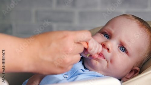 Close-up of a baby with big blue eyes being fed by an adult. The infant, wearing blue, is eating mashed food from a spoon. This candid shot captures a moment of nurturing, and early development