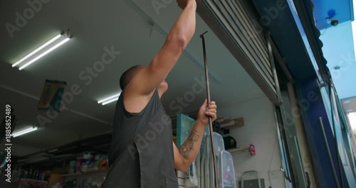 Hispanic small business hardware store owner raising metal shutter at shop entrance expressing readiness routine responsibility start of workday local entrepreneurship