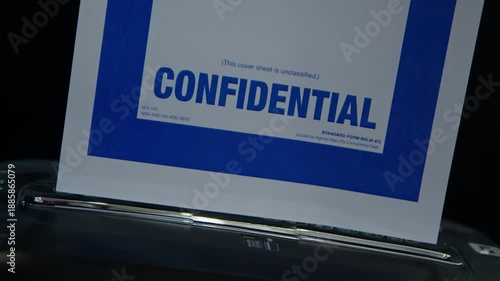 A coversheet to a confidential document is fed into a paper shredder. Subdued lighting. Suggestive of spying, espionage, or cover-up.