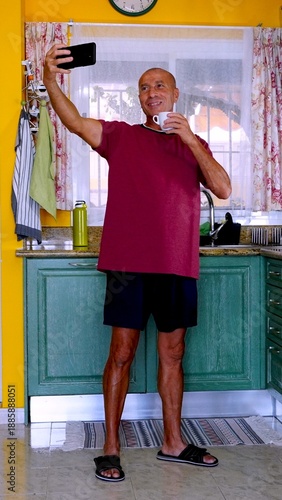 Healthy older man smiling, proudly capturing a selfie with his phone while holding a coffee cup, embodying well-being and a positive attitude in his brightly lit kitchen