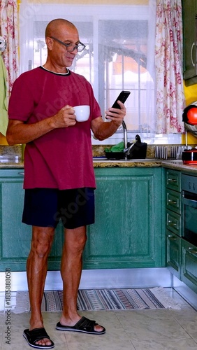 Active senior man in his kitchen standing with a coffee cup and smartphone, smiling and engaging with messages as part of a healthy, relaxed morning routine at home