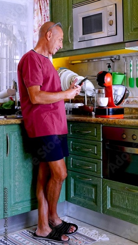 Mature man standing in a modern kitchen, casually dressed and focused on his smartphone, representing daily routine, healthy living, and digital connectivity in a domestic setting