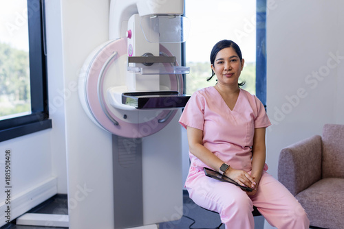 Female rad tech in pink scrubs sitting beside mammography unit with paddle and tablet, copy space