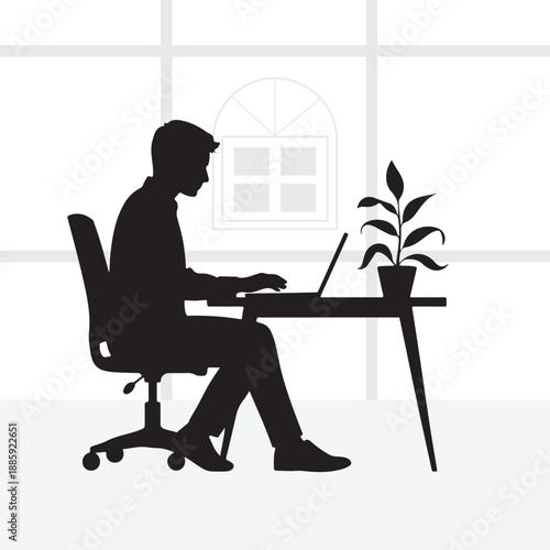 A man works remotely on his laptop at a desk with a plant and window in the background.