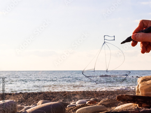 Photography Hand holding marker draws sailboat outline above pebbly beach and sea horizon
