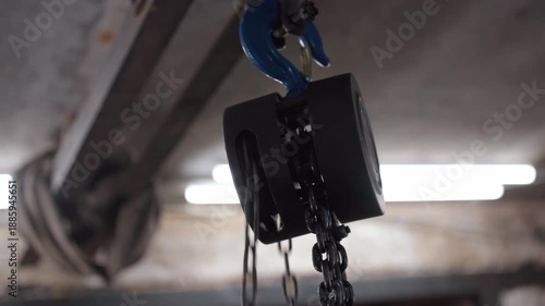 Close-up of a black chain hoist pulley system slowly moving in an industrial warehouse. The focus is on the mechanism lifting heavy equipment in a factory or construction site