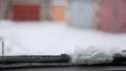 Close-up of a car's windshield wiper blade sweeping back and forth, cleaning away fresh snow and ice from the glass during a winter storm for better visibility while driving