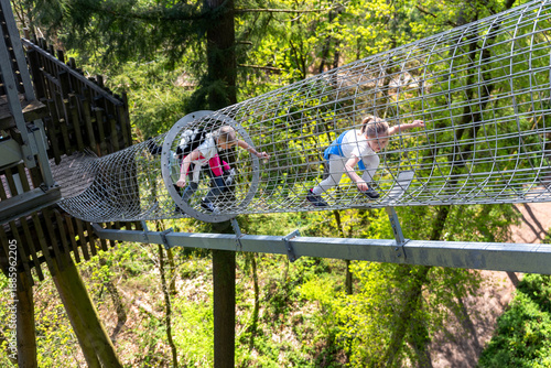 Two cute little funny blond girl friends enjoy having fun walk metal net tunnel playground in treetop adventure amusement park Saarland. Children outdoors nature extreme sport activities concept