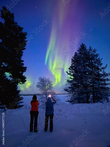 Two girls standing in front of Northern Lights and taking pictures of this majestic natural phenomenon during ice cold polar night close to the cities of Inari and Ivalo in  Lapland, Finland