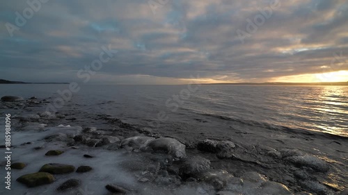 Rocky lakeshore with patches of ice and snow at sunset, dark rippling water under a dramatic cloudy sky, cold winter evening landscape with soft golden light on the horizon over the distant coast