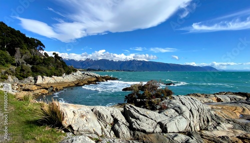 Rocky coastline with vibrant blue water under a sunny sky and fluffy clouds, with mountains in the distance