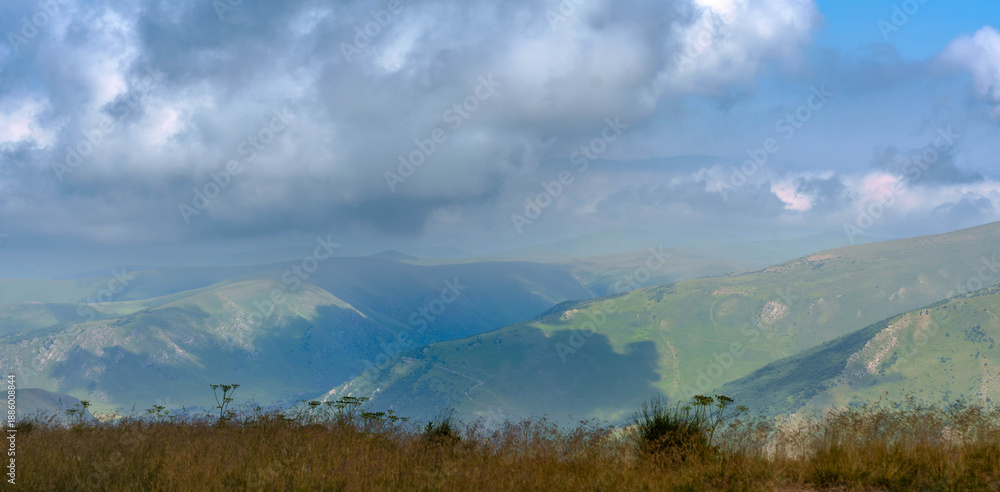 Fototapeta premium Wide view of green hills and mountains under cloudy blue sky.Expansive landscape with layered green hills and distant mountains under a cloudy blue sky. Wild plants in foreground