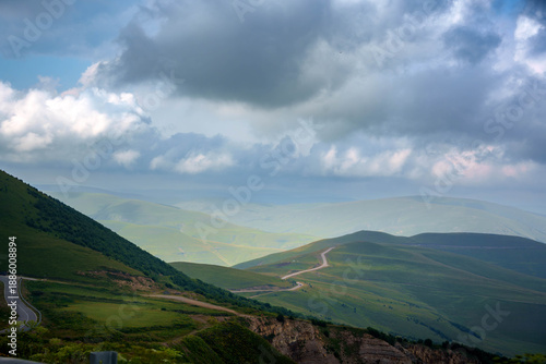 Winding mountain road through green hills in the Caucasus region.Scenic view of a winding dirt road cutting through green hills and mountains in the Caucasus region. Misty background and dramatic sky