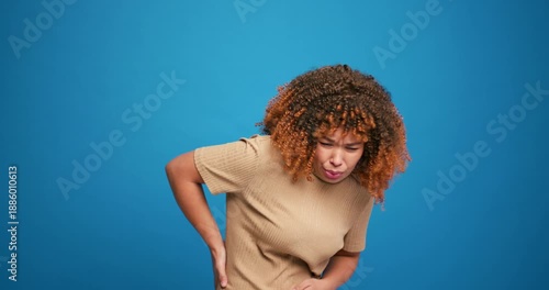 Sad, sick woman with curly hair having strong pain in beck and in across isolated on blue background