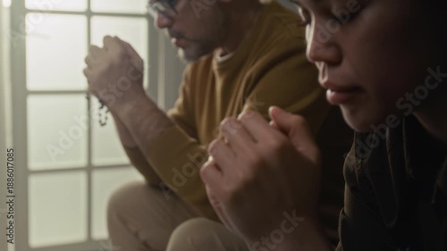 Close-up rack focus view of young woman whispering prayer with eyes closed while praying next to mature spiritual mentor holding rosary