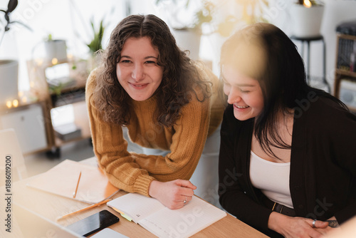 Two young women collaborate on a project at a desk, reviewing notes and smiling.