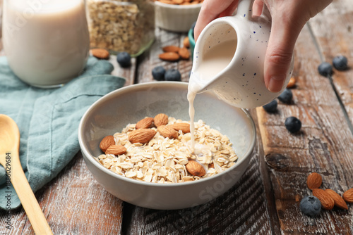 Foto Woman pouring almond milk into bowl with oat flakes and nuts at wooden table, cl