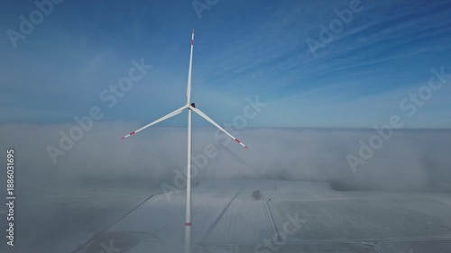 Wind turbine spins against a backdrop of blue sky and fog, showcasing the transition from clear visibility to a misty landscape in a rural setting