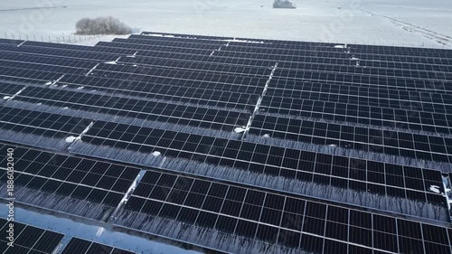Aerial view of solar panels covered in snow, showcasing rows of photovoltaic cells in a winter landscape with a distant tree line and open fields