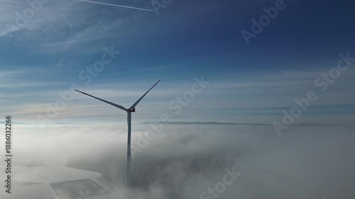 Wind turbine generating electricity in thick fog above landscape with clouds and blue sky