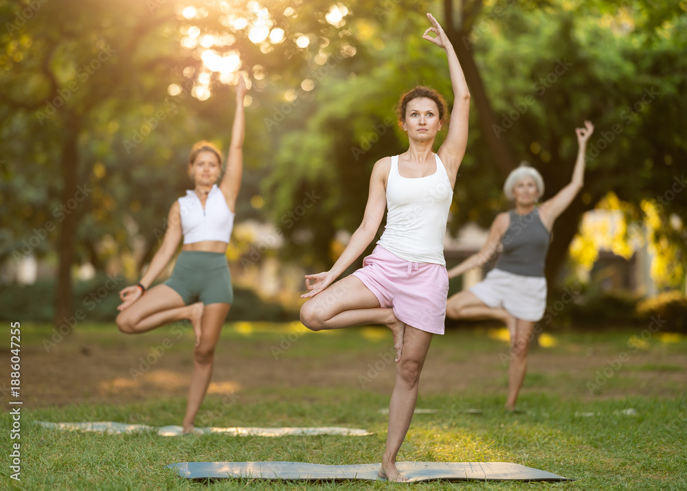 Fototapeta premium Sporty middle-aged woman practicing Vrksasana balancing asana, or Tree Pose, in sunlit green city park during group yoga session. Healthy active lifestyle concept