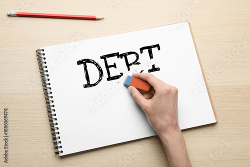 Photos Woman erasing word Debt in notebook at wooden table, top view