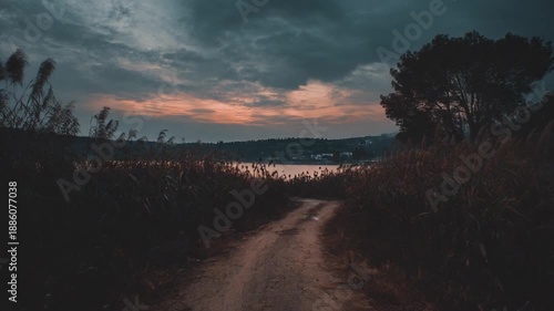 A dirt road leads through tall grass towards a tranquil lake and a dusky sky at twilight