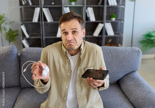Charger smartphone phone cable has charging problem. A puzzled confused man inspects a faulty adapter at home, in frustration, while the device will not power up. Clear failure needs quick repair.