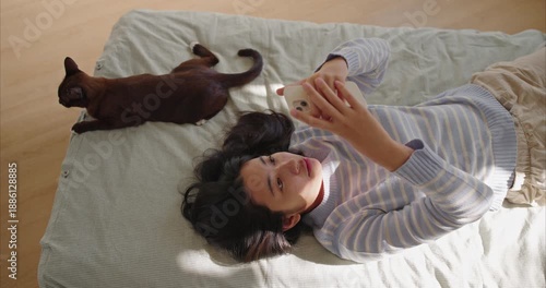 young Asian woman lies on a tidy bed using a smartphone as a brown cat lounges nearby. Morning light casts warm highlights on pale wood floor and pastel bedding. top view