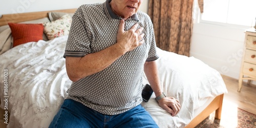 Photos Elderly man sitting on bed, hand on chest, appearing unwell
