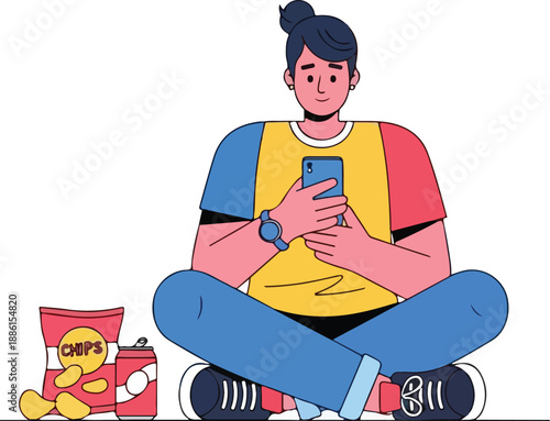 A young man sits cross-legged on the floor, engrossed in his smartphone, snacking on chips beside him.