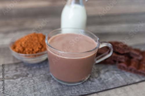 Cup of hot cocoa with milk bottle, chocolate pieces and cocoa powder on rustic wooden table. 