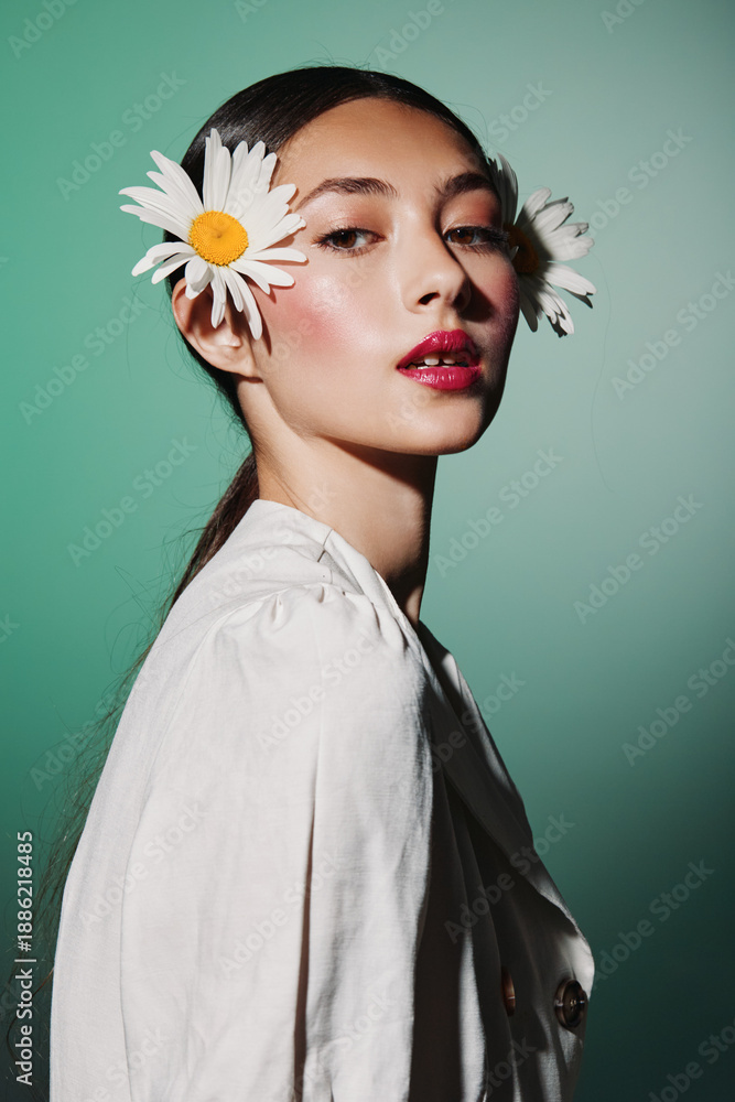 Fototapeta premium Portrait of a stylish woman with daisies in her hair wearing light clothes on green background. High fashion beauty concept with natural floral accessories and makeup.