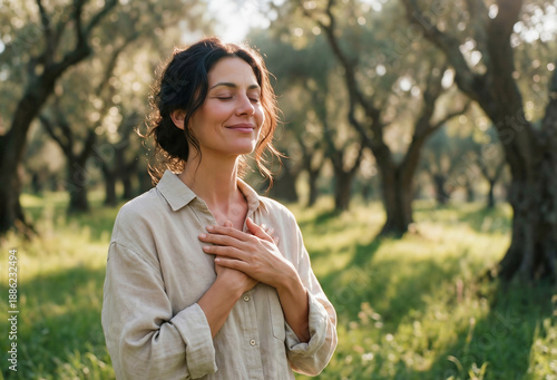 Smiling woman with closed eyes and hands on her chest in a sunlit olive grove, embodying gratitude, peace, and mindfulness