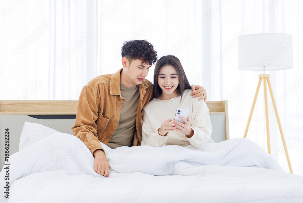 Fototapeta premium Asian young couple love happy sharing using mobile smartphone sitting lying on bed in white bedroom at home together boyfriend girlfriend relationship lifestyle relax beautiful romantic Valentine day.