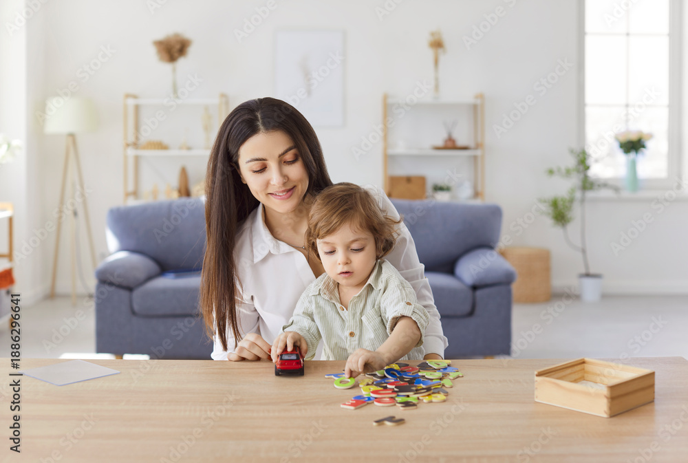 Naklejka premium Smiling young mother sitting at desk and playing with her little son holding toy car at home. Parent and kid making words of letters or learning alphabet during educational activity in living room.