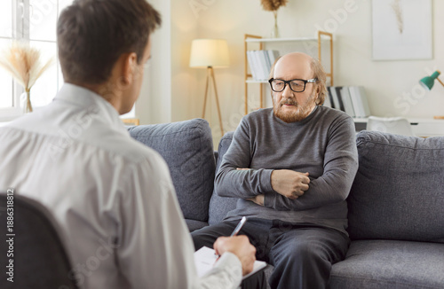 Foto Male young professional psychologist doctor making notes consulting senior elderly man patient sitting on sofa during therapy session in office
