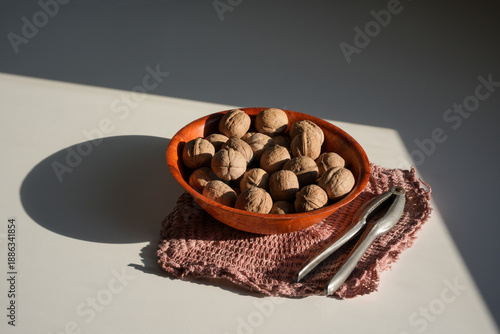 A wooden bowl filled with whole walnuts in their shells, resting on a textured pink/brown placemat. A metal nutcracker is placed next to the bowl on a white table.
