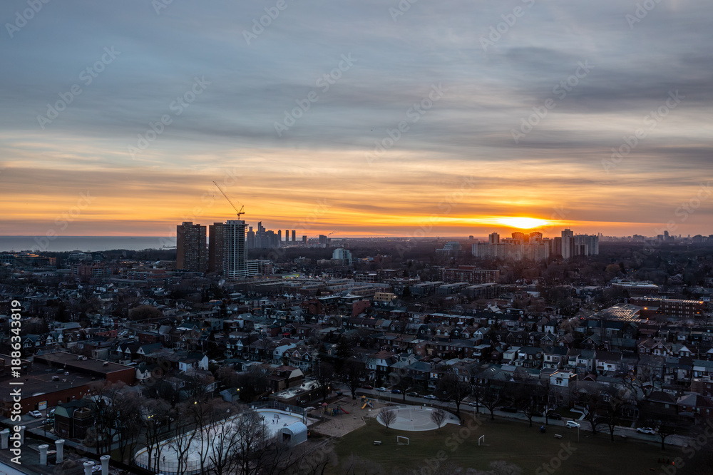 Fototapeta premium Toronto cityscape showing residential homes and high rise buildings illuminated by the setting sun casting warm orange hues across the sky above an outdoor skating rink and park