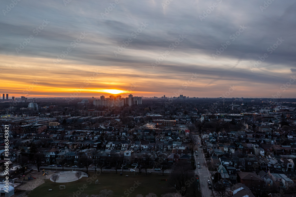 Fototapeta premium Toronto skyline and sprawling residential neighborhoods bathed in warm orange sunset light, dramatic clouds over urban and suburban development stretching to the horizon at dusk