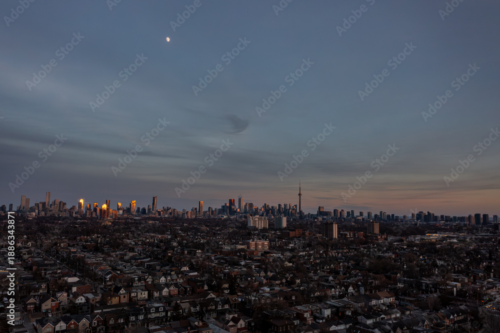 Fototapeta premium Toronto cityscape featuring the iconic skyline and a residential neighborhood, with the moon visible in the twilight sky and buildings reflecting warm golden light during a serene evening