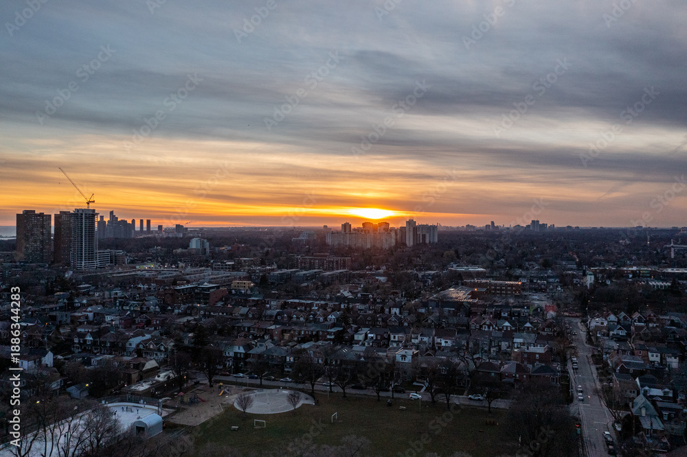 Fototapeta premium Toronto cityscape featuring residential neighborhoods and an urban park, bathed in the warm glow of a setting sun painting the sky with orange and yellow hues, highlighting the vast city expansion