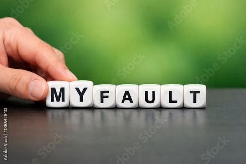 Personal Responsibility: A close-up shot of a hand arranging letter dice to spell MY FAULT, against a blurred verdant background, embodies culpability.
